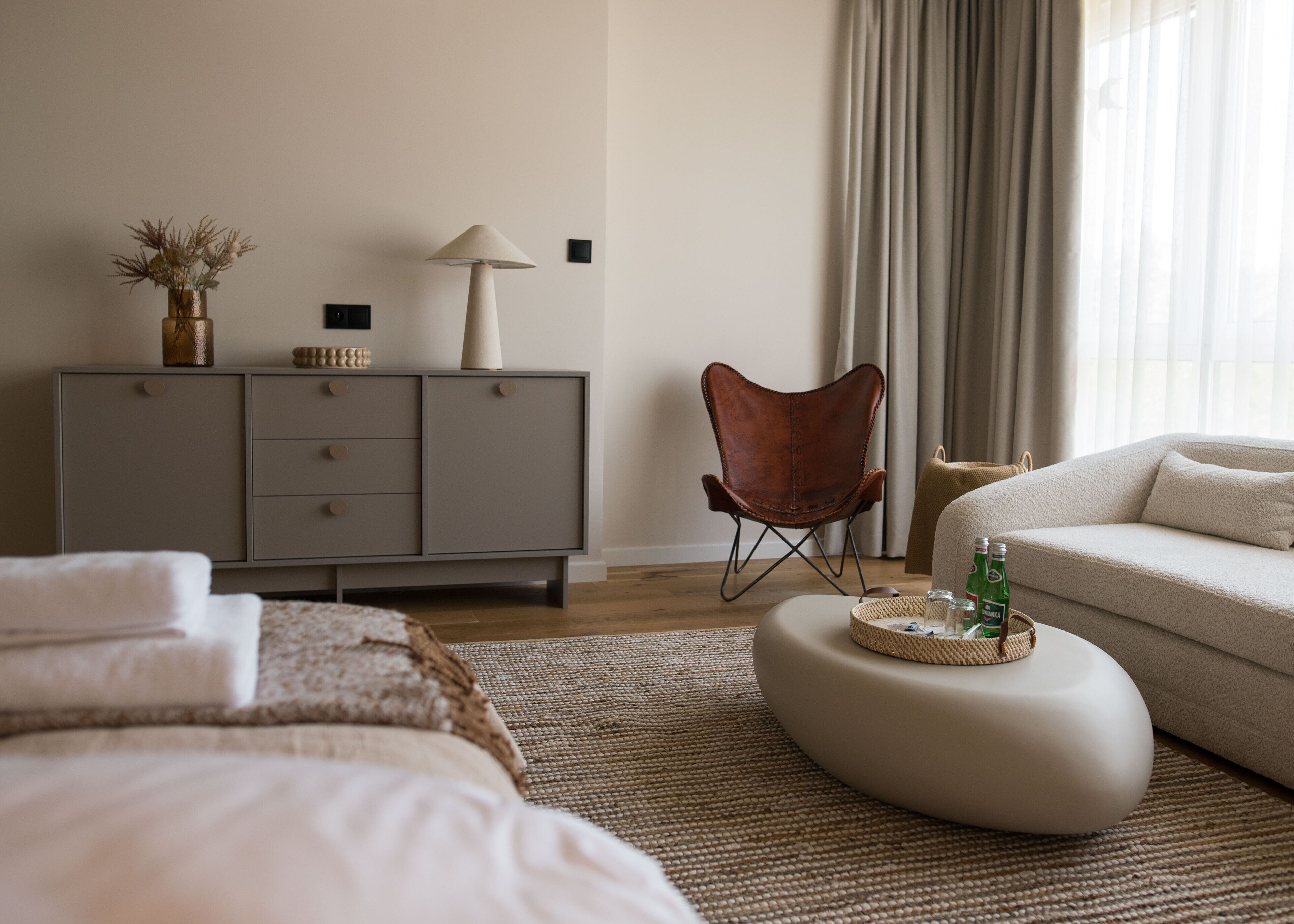 A modern bedroom with a bed, a grey dresser, a leather chair, a sofa, and a round ottoman with bottles.