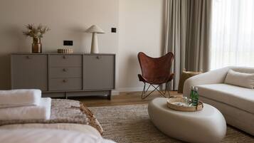 A modern bedroom with a bed, a grey dresser, a leather chair, a sofa, and a round ottoman with bottles.