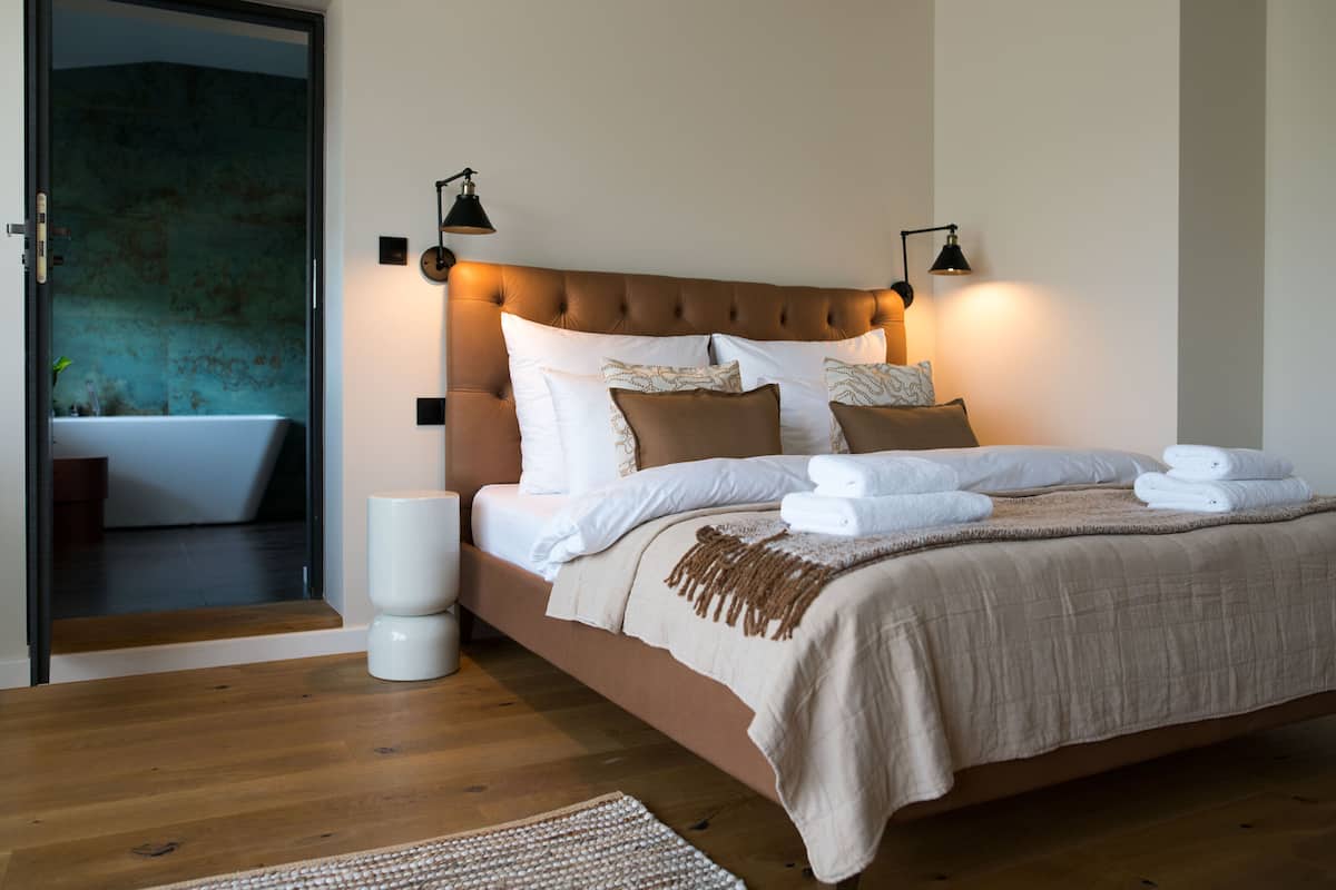 A modern bedroom with a tufted headboard, a large bed with multiple pillows, a bedside lamp, and a neatly made bed with folded towels.