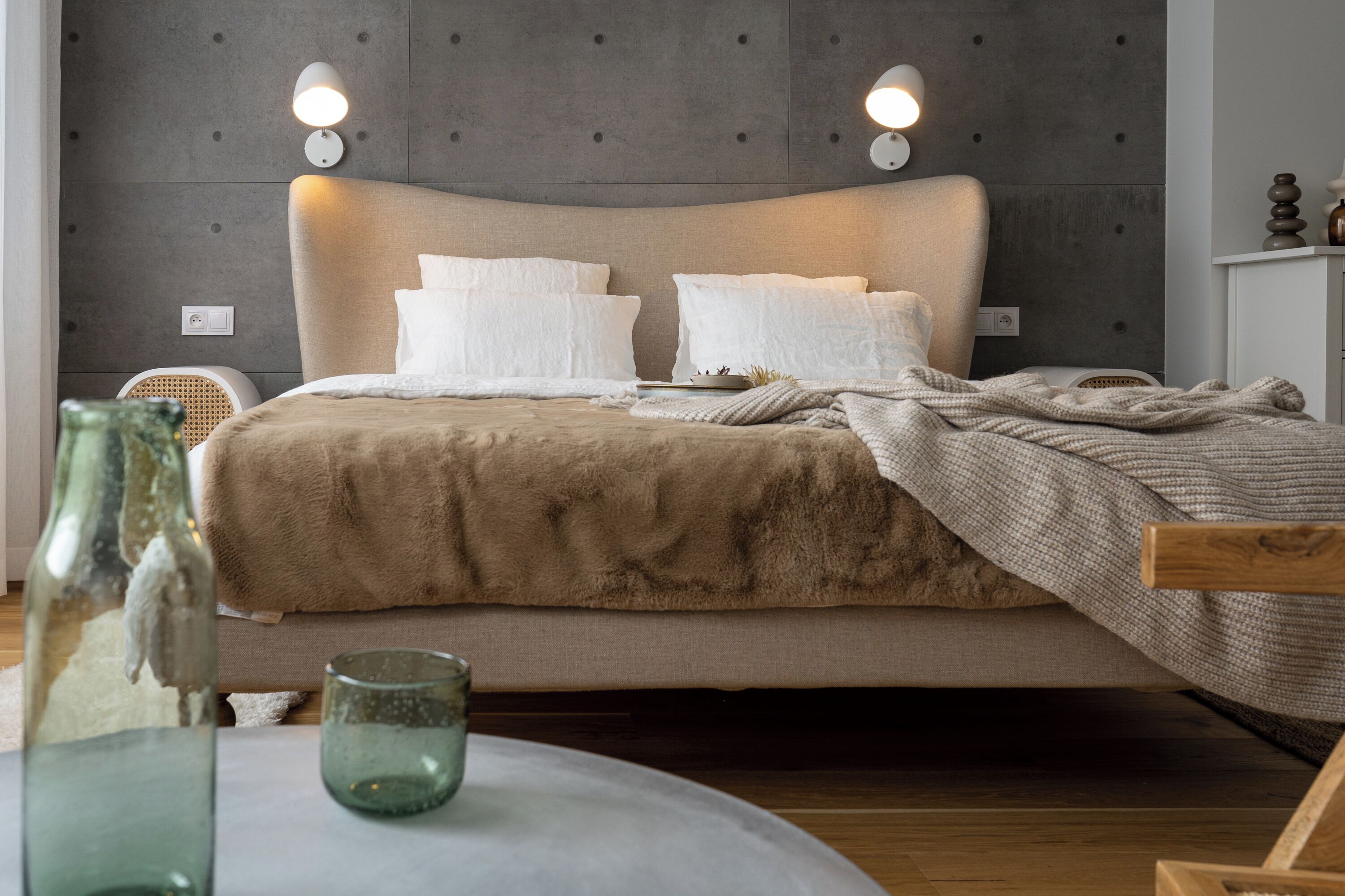 A bedroom with a large bed, two wall-mounted lights, a bedside table with a glass container, and a chair.