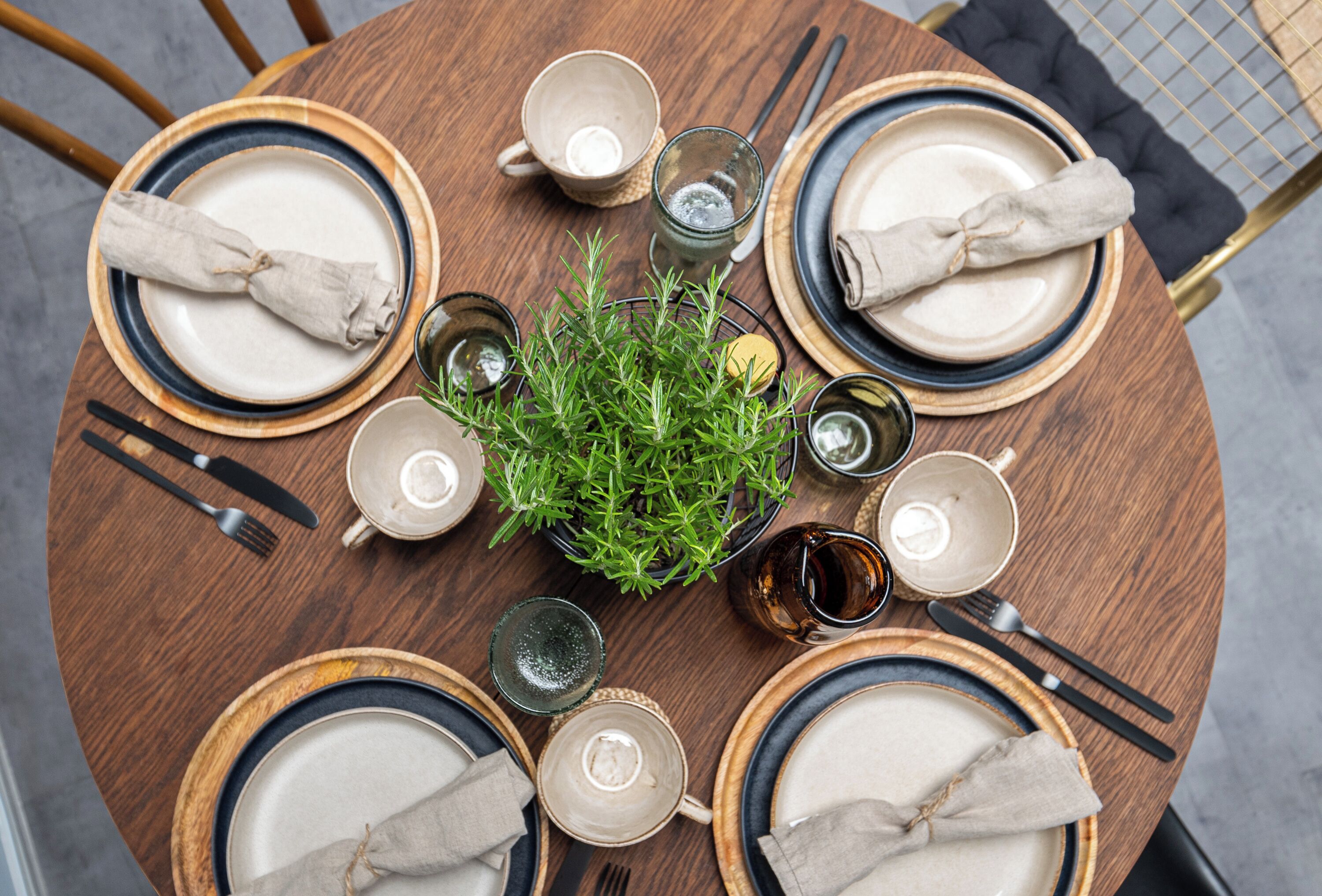 A round wooden table set for four with plates, cutlery, glasses, and a central plant.