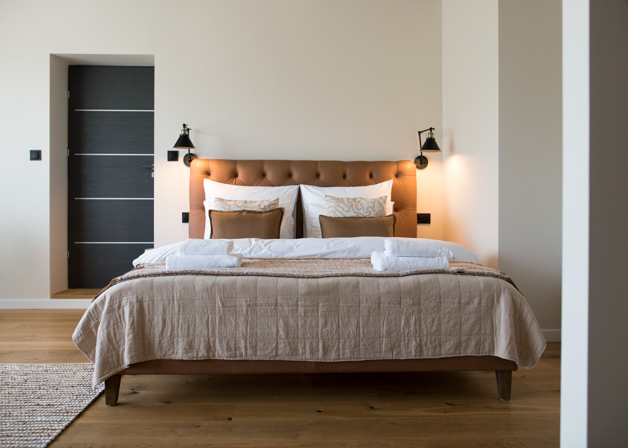 A neatly made bed with a beige bedspread and pillows, a dark door in the background, and two wall-mounted lamps on either side of the bed.