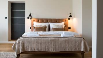 A neatly made bed with a beige bedspread and pillows, a dark door in the background, and two wall-mounted lamps on either side of the bed.