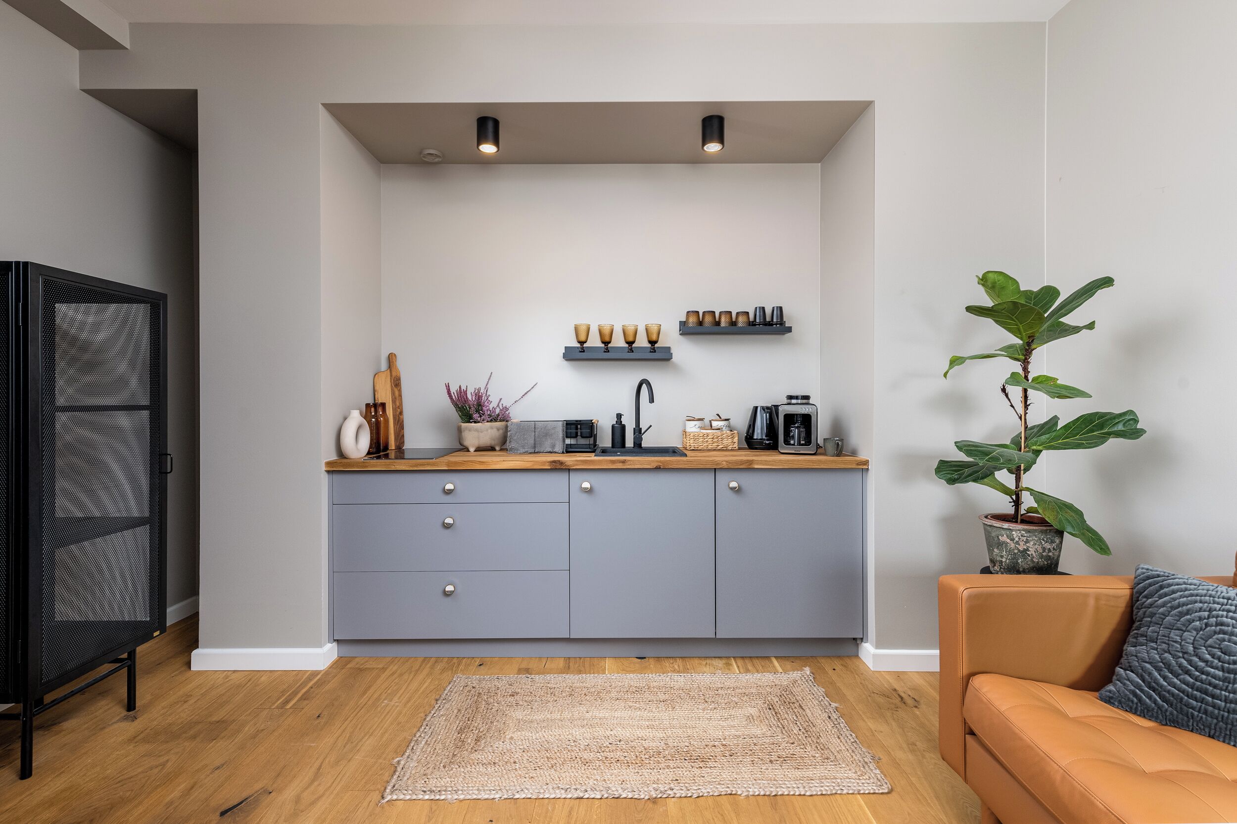 Una cocina compacta con armarios grises, encimera de madera y un lavamanos. Hay estantes sobre la encimera con tazas y una planta. En la zona de estar, hay un sofá marrón y una planta grande en maceta.
