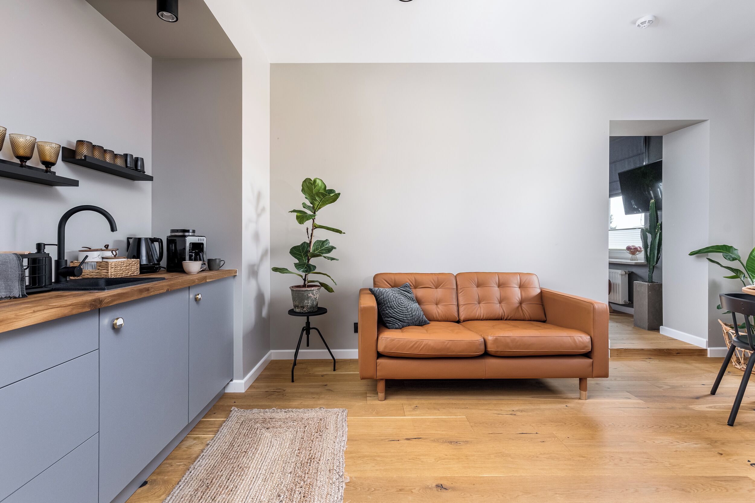 Una cocina moderna con gabinetes grises, encimera de madera y grifo negro. Hay un sofá de cuero marrón con un cojín y una planta en maceta a su lado.
