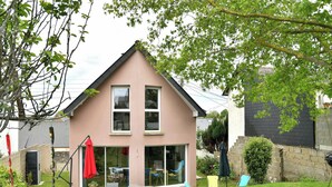 Une maison à deux étages, dotée d’un toit à deux versants, d’une porte d’entrée et d’une porte-fenêtre coulissante donnant sur un patio aménagé avec du mobilier d’extérieur.