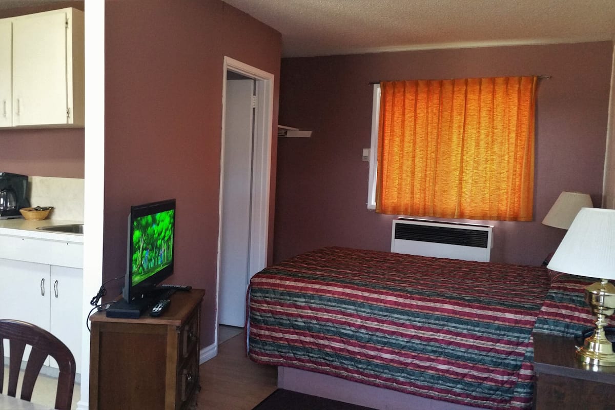 Un dormitorio con cama, televisor, ventana con cortinas, escritorio con lámpara y kitchenette con microondas.