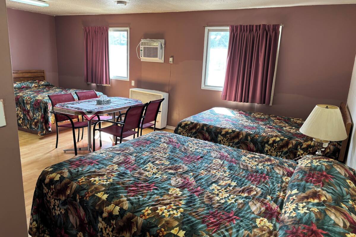 Una habitación con dos camas, una mesa con sillas, una lámpara y una ventana con cortinas.