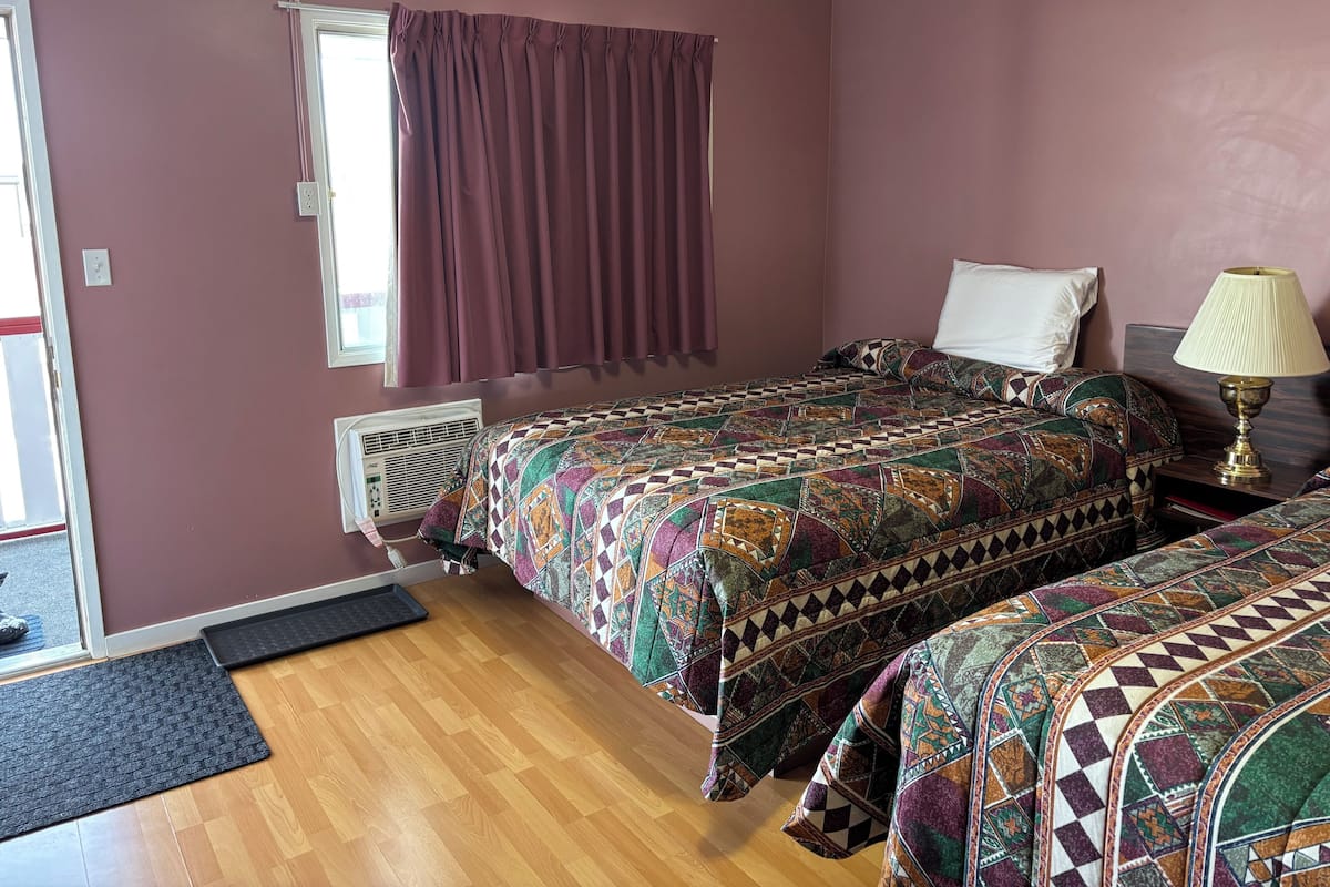 Una habitación con dos camas, una ventana con cortinas, un radiador y una lámpara de noche.