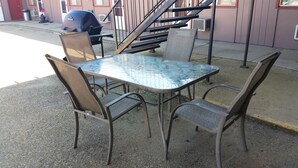 An outdoor seating area with a metal table and chairs, a barbecue grill, and a staircase leading to an upper level. - Lamplighter Viking Motel (Viking)