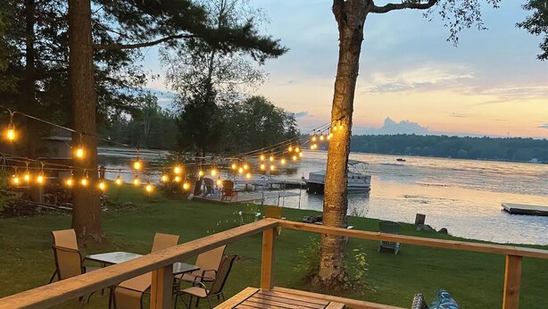 A deck with outdoor seating overlooking a lake, string lights, and a boat in the distance. - Waterfront Cottage ~ Fireplace ~ Kawartha (Lakefield)