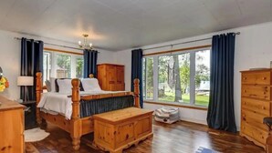 A bedroom with a wooden bed, a wooden chest of drawers, and a large window with curtains. - Waterfront Cottage ~ Fireplace ~ Kawartha (Lakefield)