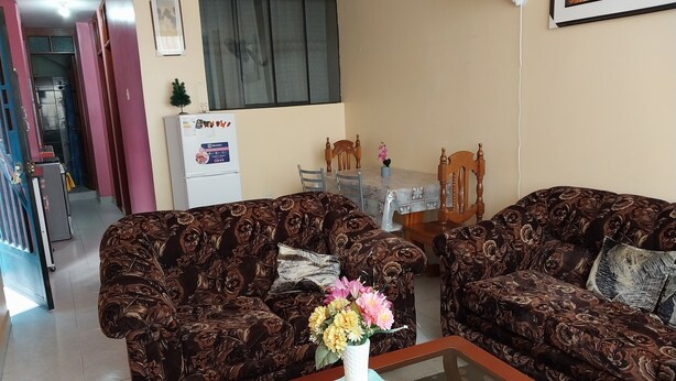 A living room with two patterned sofas, a coffee table with a vase of flowers, a dining table with chairs, and a refrigerator.
