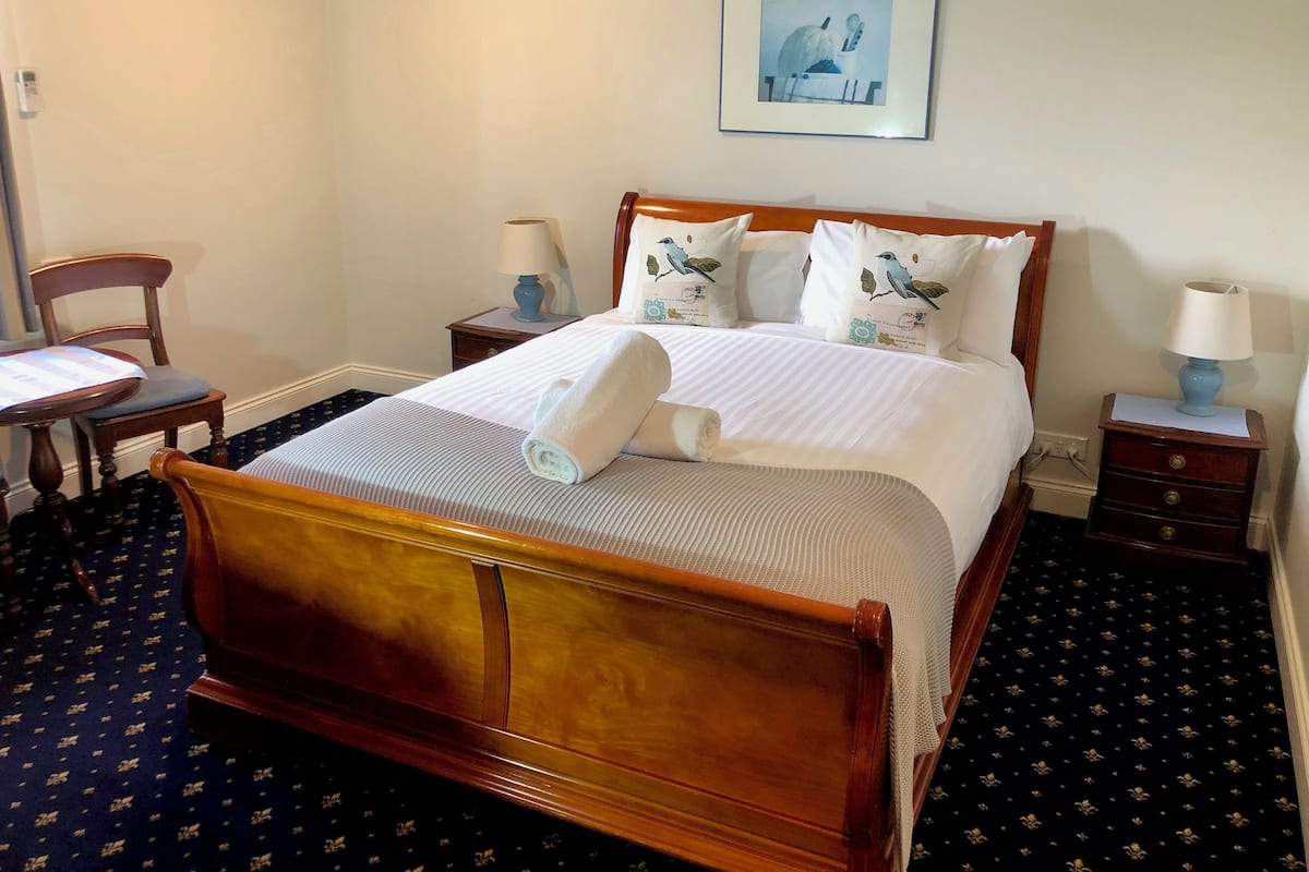 A neatly made bed with a rolled towel, a wooden nightstand with a lamp, a chair, and a framed picture on the wall.