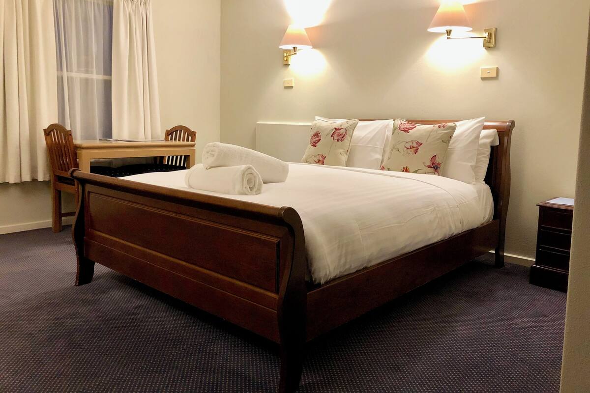 A hotel room with a wooden bed, a desk, and a chair. There are two wall-mounted lamps and a window with curtains.