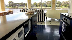 A kitchen with a large island, a dining area with chairs, and a view of a river and greenery. - Douglas Riverside (Port Fairy)