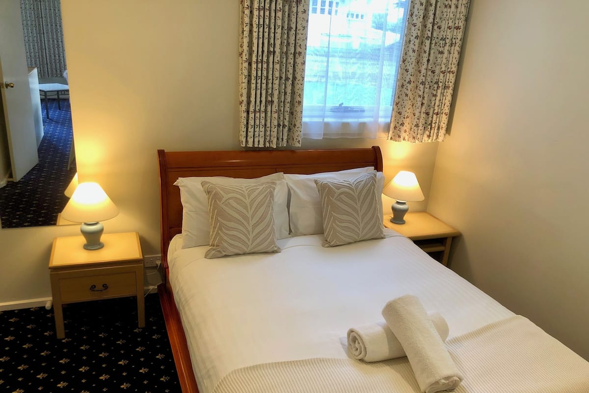 A neatly made bed with two pillows, a rolled towel, and two bedside lamps.