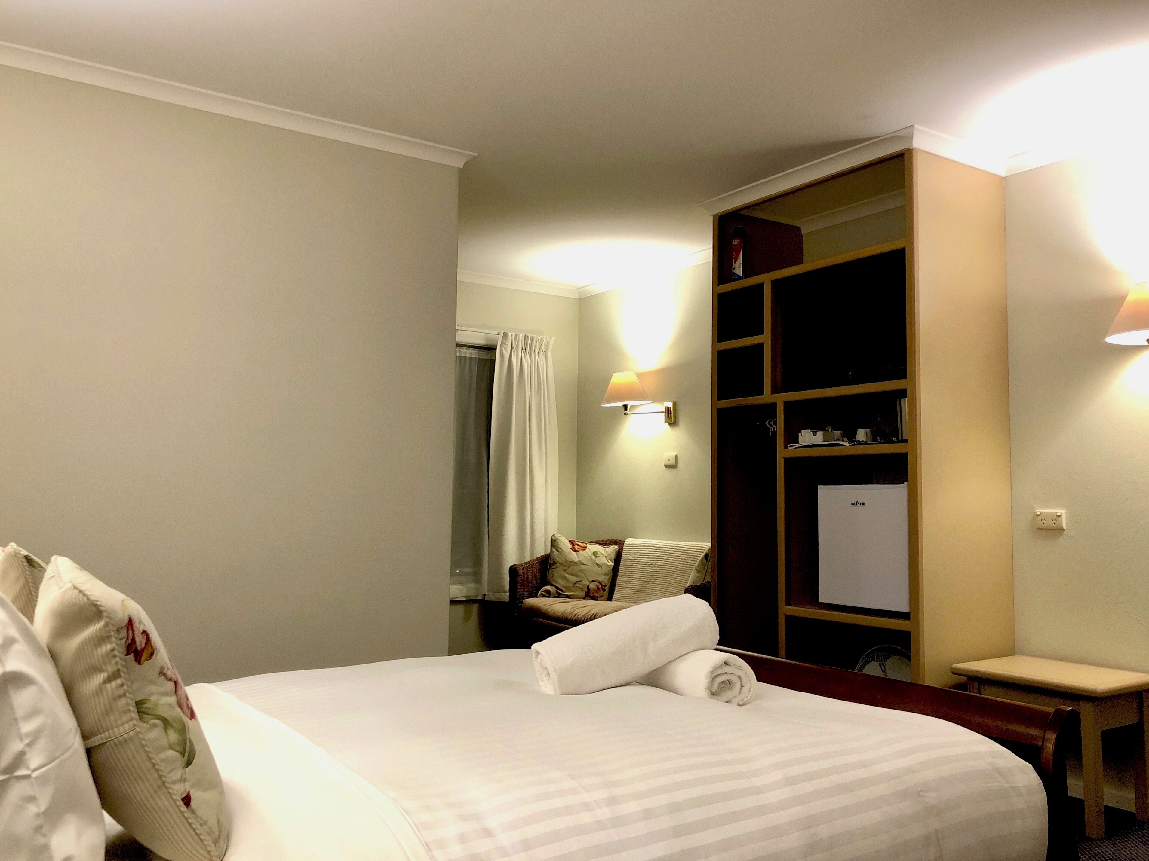 Una habitación de hotel con una cama, un armario de madera, una mesita y una lámpara de pared.