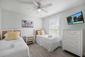 A hotel room with two beds, a ceiling fan, a TV, and a painting on the wall.