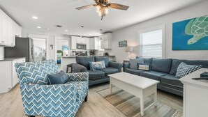 A modern living room with a sofa, armchair, and coffee table. There is a ceiling fan, a painting of a turtle, and a kitchen in the background. - Family Tides A – AMI Retreat w/ Pool, Rooftop Deck & Gulf/Bay Views Steps To Beach (Bradenton Beach)