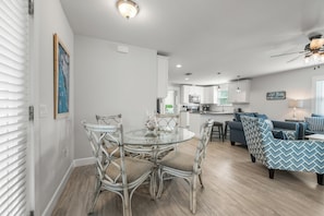 Un salón espacioso con una mesa de comedor de vidrio rodeada de sillas de mimbre, un sofá azul con cojines estampados y una cocina al fondo.