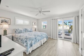 Un dormitorio con cama, ventilador de techo, ventana con persianas, mesita de noche con lámpara y vista al balcón exterior.