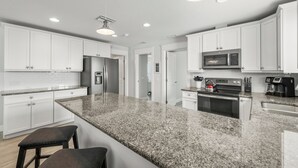 A modern kitchen with white cabinets, granite countertops, stainless steel appliances, and a central island with stools. - Family Tides A – AMI Retreat w/ Pool, Rooftop Deck & Gulf/Bay Views Steps To Beach (Bradenton Beach)
