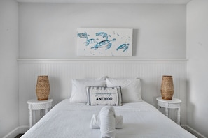 A bedroom with a white bed, two bedside tables with woven baskets, a wall-mounted artwork of sea turtles, and a pillow with the word 'ANCHOR'. - Family Tides B – AMI Retreat w/ Pool, Rooftop Deck & Gulf/Bay Views Steps To Beach (Bradenton Beach)