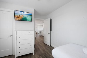 A bedroom with a bed, a dresser, a television mounted on the wall, and a view of a beach through the window.