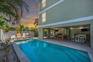 A pool area with lounge chairs and a table, surrounded by a building with a flat roof and a covered patio. - Family Tides B – AMI Retreat w/ Pool, Rooftop Deck & Gulf/Bay Views Steps To Beach (Bradenton Beach)