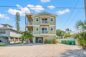 A two-story building with a balcony on the second floor, a covered entryway, and a sandy driveway. - Family Tides B – AMI Retreat w/ Pool, Rooftop Deck & Gulf/Bay Views Steps To Beach (Bradenton Beach)