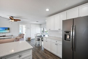 A modern kitchen with white cabinets, a stainless steel refrigerator, and a dining area with a table and chairs. - Family Tides B – AMI Retreat w/ Pool, Rooftop Deck & Gulf/Bay Views Steps To Beach (Bradenton Beach)