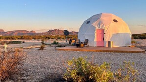 A geodesic dome structure with a pink door, surrounded by outdoor seating and a satellite dish, set against a desert landscape with mountains in the background. - Southern Boho inspired Geodesic Dome nestled up next to Big Bend National Park. (Terlingua)