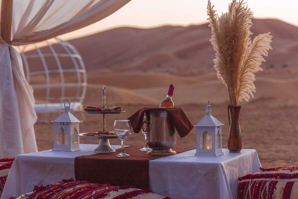 Un montaje para comer en el desierto, con una mesa dispuesta para dos personas, que incluye postre, una jarra, vasos y faroles.