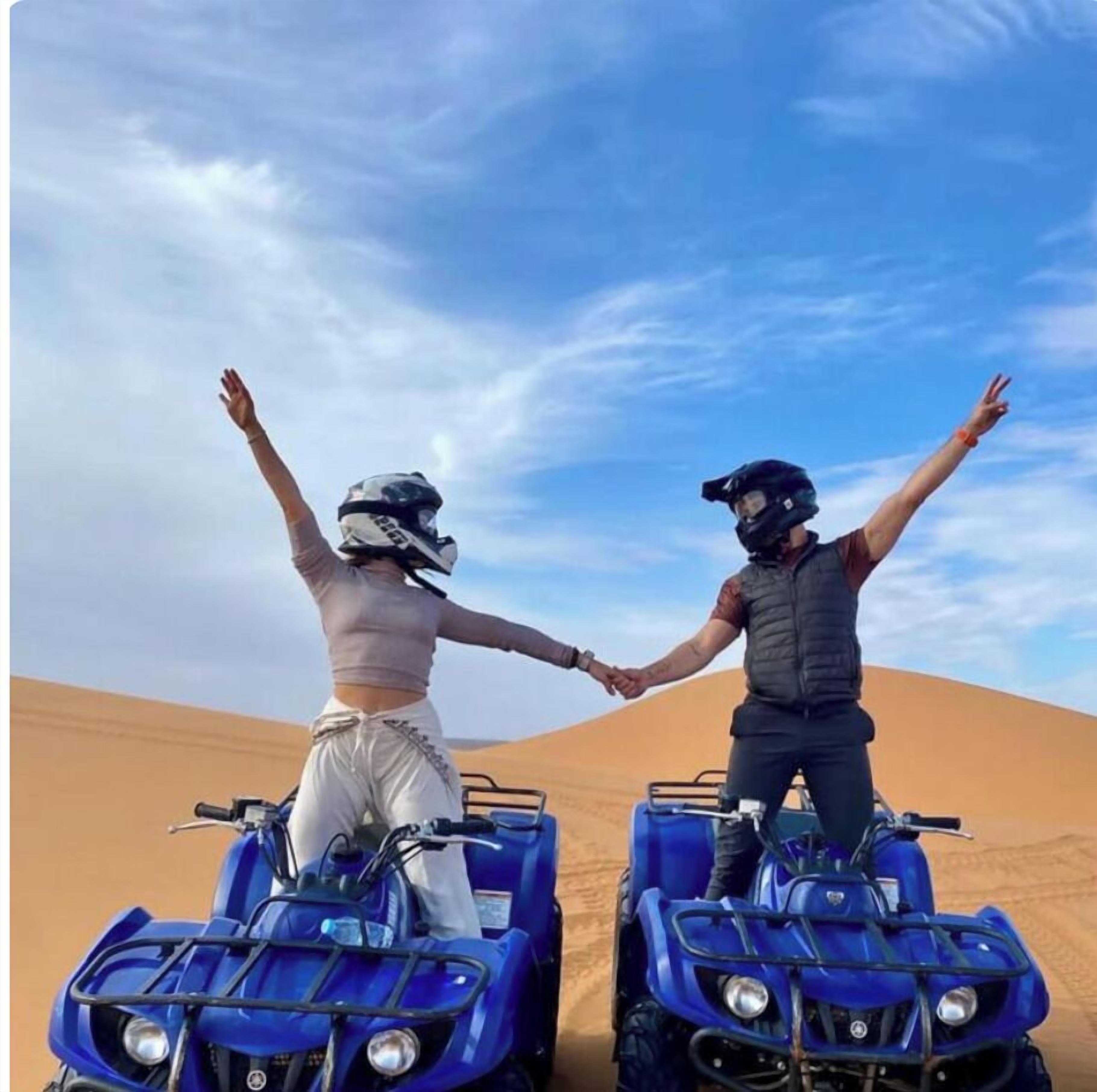 Two people on ATVs in a desert with sand dunes, holding hands.