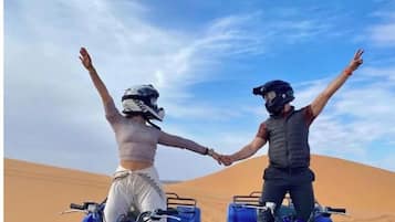 Two people on ATVs in a desert with sand dunes, holding hands.
