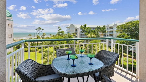A balcony with wicker chairs and a glass table, overlooking a beach and a resort complex. - 3402 Windsor @ Palmetto Dunes | Oceanfront | Steps to Beach | Pool | Stunning Views (Hilton Head Island)
