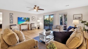 A spacious living room with a ceiling fan, a large TV, and a view of the outdoors through a sliding glass door. - 542 Queen's Grant @ Palmetto Dunes | Dog-Friendly | Pool | Walk to Beach | Golf (Hilton Head Island)