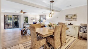 A spacious living area with a wooden dining table, wicker chairs, a ceiling fan, and a view through glass doors to an outdoor patio. - 542 Queen's Grant @ Palmetto Dunes | Dog-Friendly | Pool | Walk to Beach | Golf (Hilton Head Island)