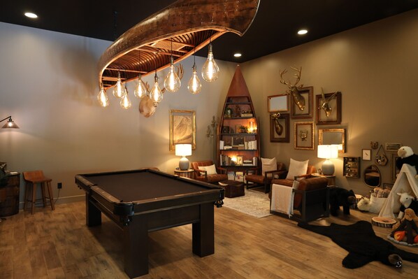 A pool table in a room with a wooden floor, a bookshelf, and a lamp.