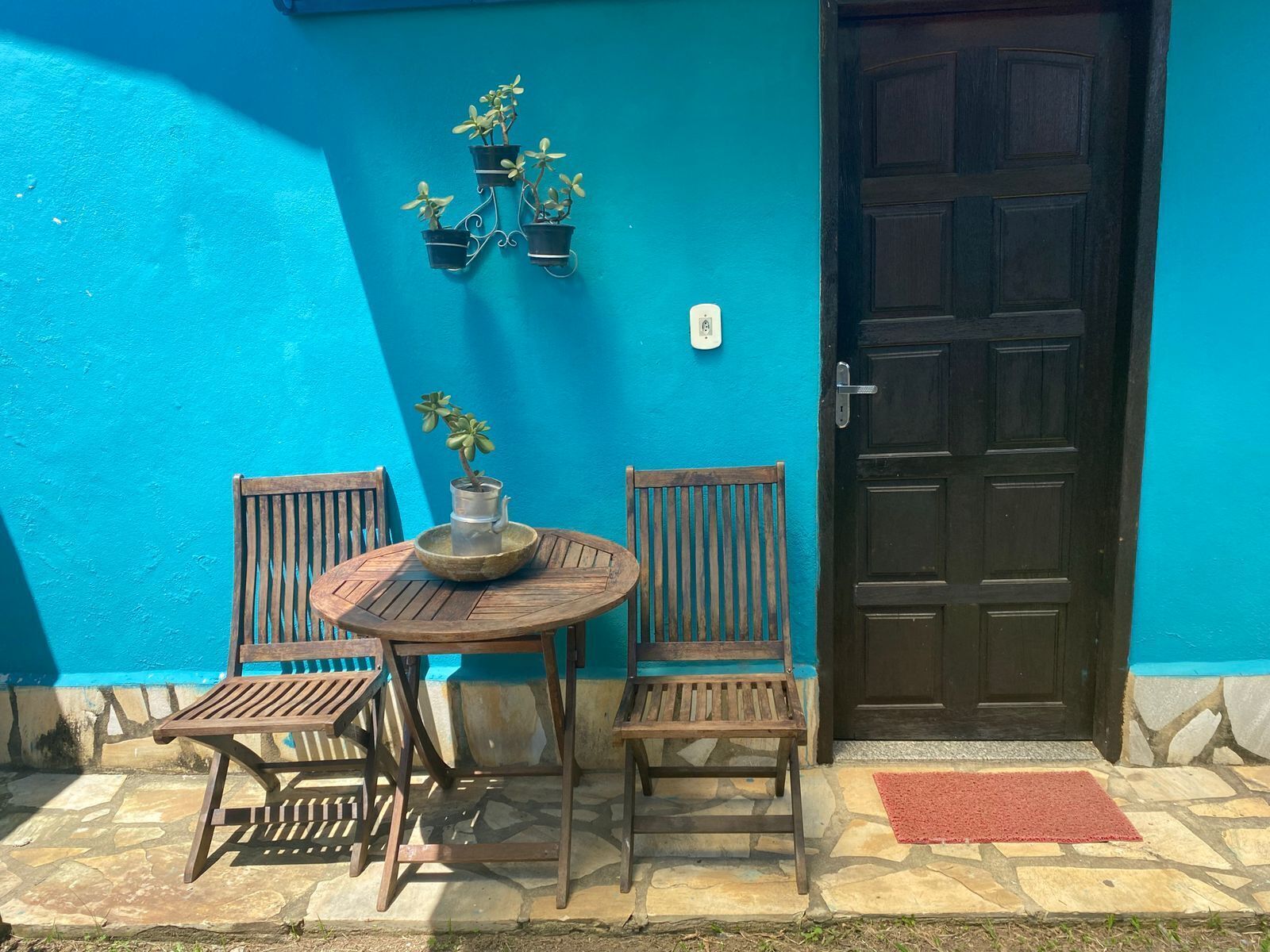 Een houten tafel met stoelen en een bloempot voor een blauwe muur met een deur.