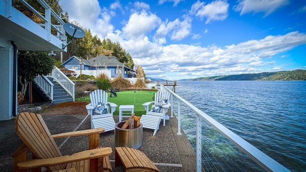 En træterrasse med Adirondack-stole, bålsted og udsigt til sø og fjerne huse.