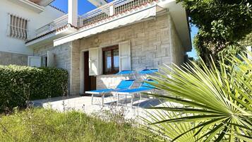 Un edificio a due piani con facciata in pietra, un balcone con ringhiere bianche e un patio con poltrone da relax blu.