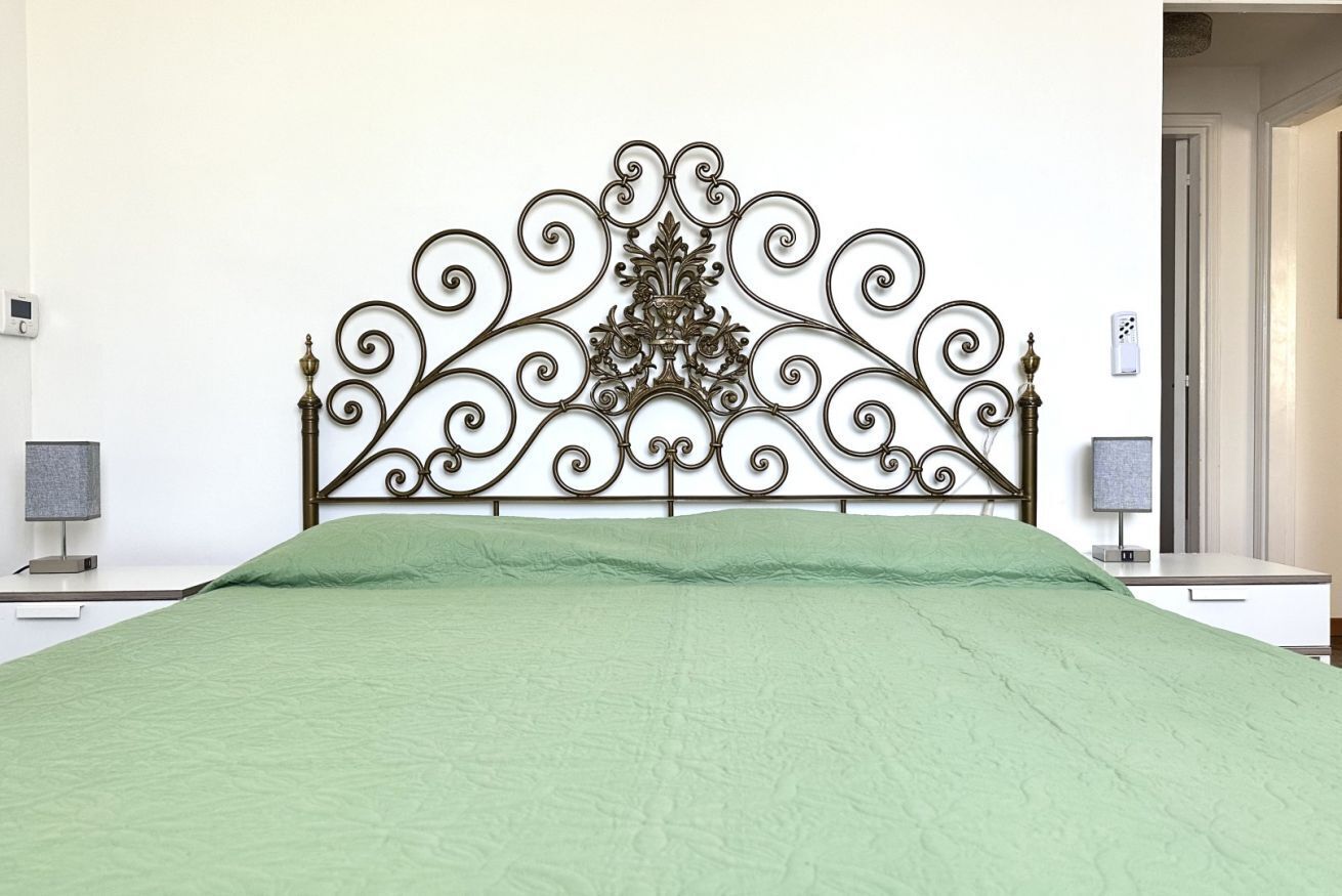 Un letto con copriletto verde, una testiera decorativa in metallo, comodini con lampade e una parete bianca.