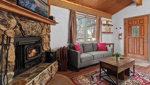 Ein gemütliches Wohnzimmer mit Kamin aus Stein, einem grauen Sofa mit roten Kissen, einem Couchtisch aus Holz und einem großen Fenster mit Vorhängen.