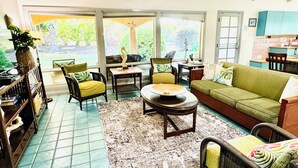 A spacious living area with a green sofa, two armchairs, a coffee table, and a bookshelf. Large windows provide a view of the outdoors.