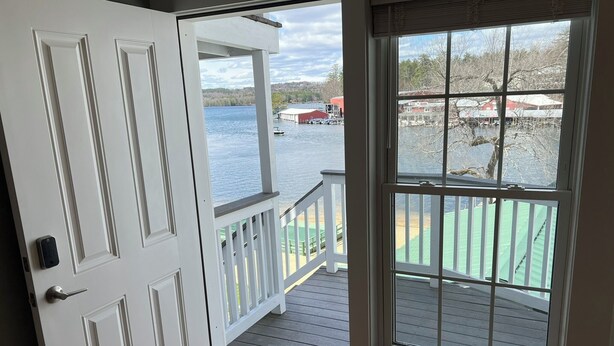 A view from a room with a door leading to a deck overlooking a body of water, with a view of buildings and trees in the distance. - Spectacular Sunset Suite (Laconia)