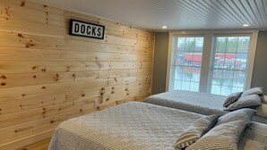 A room with two beds, a wooden wall with a 'DOCKS' sign, and a window with a view of a dock and boats. - Spectacular Sunset Suite (Laconia)