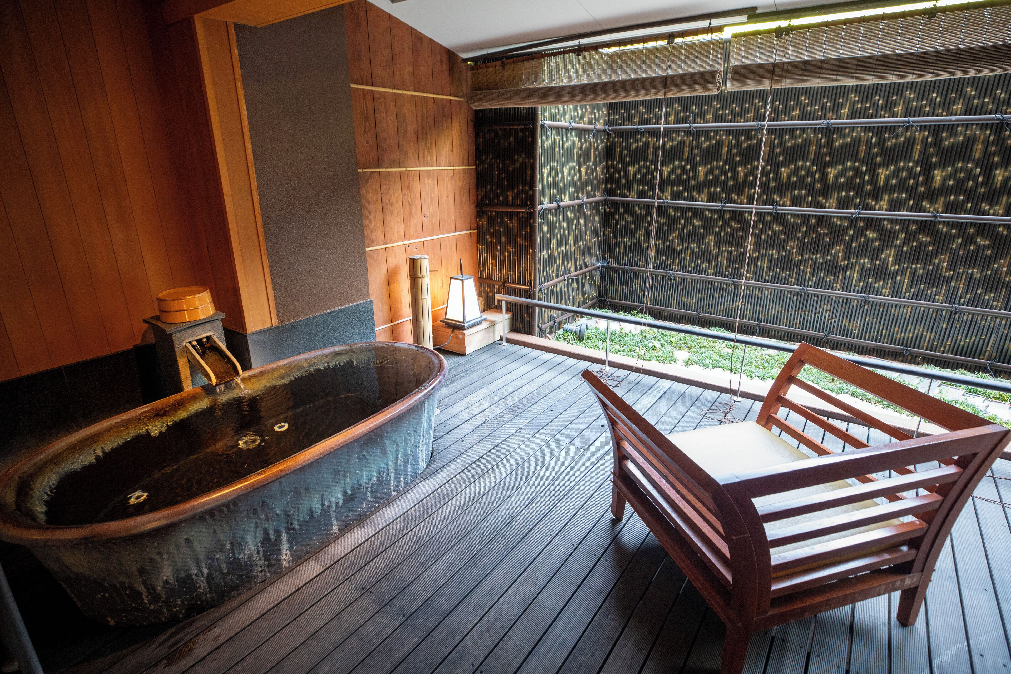 A wooden deck with a large bathtub, a wooden chair, and a view through a bamboo screen.