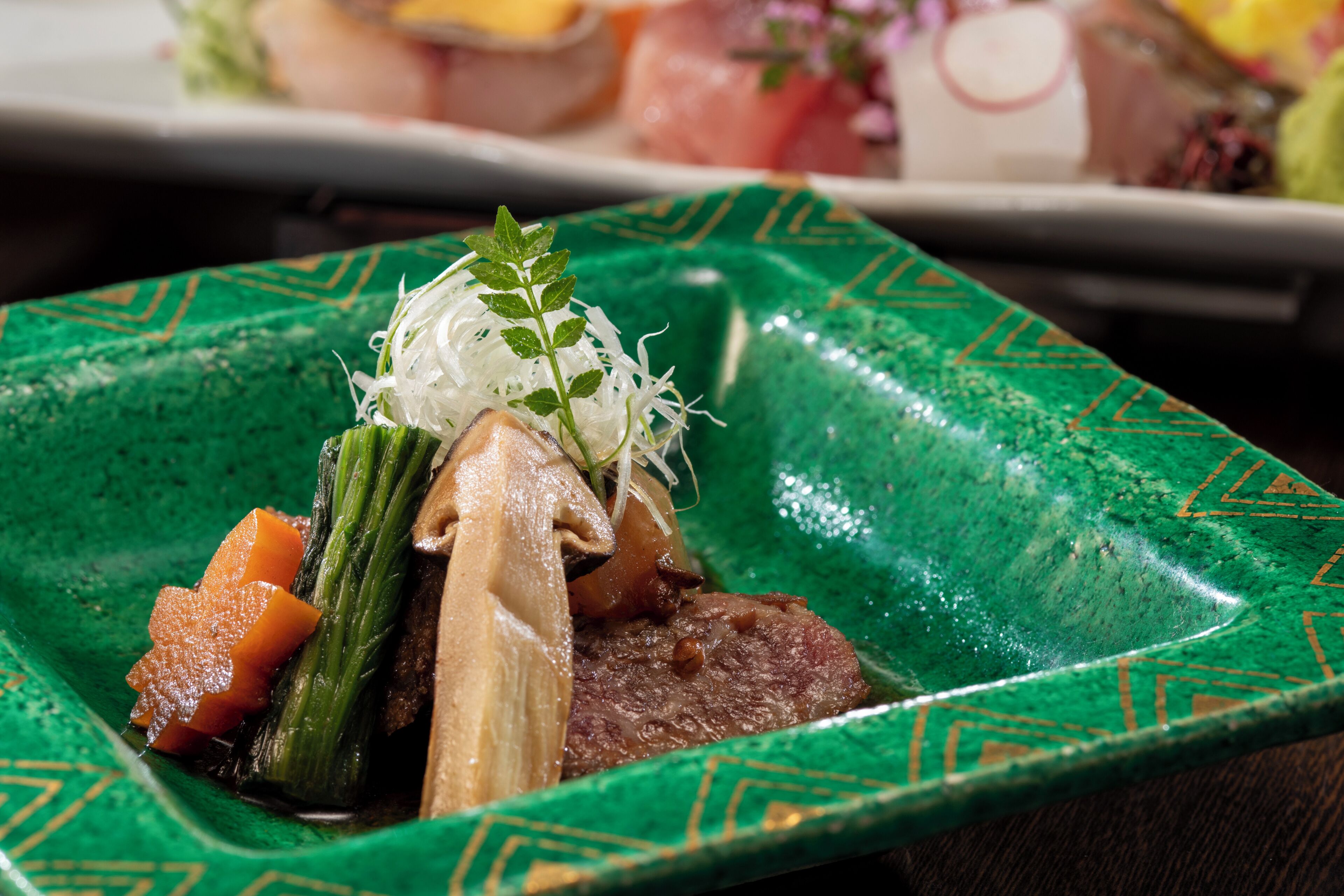 A square plate with a green pattern, containing a dish with meat, vegetables, and a garnish.
